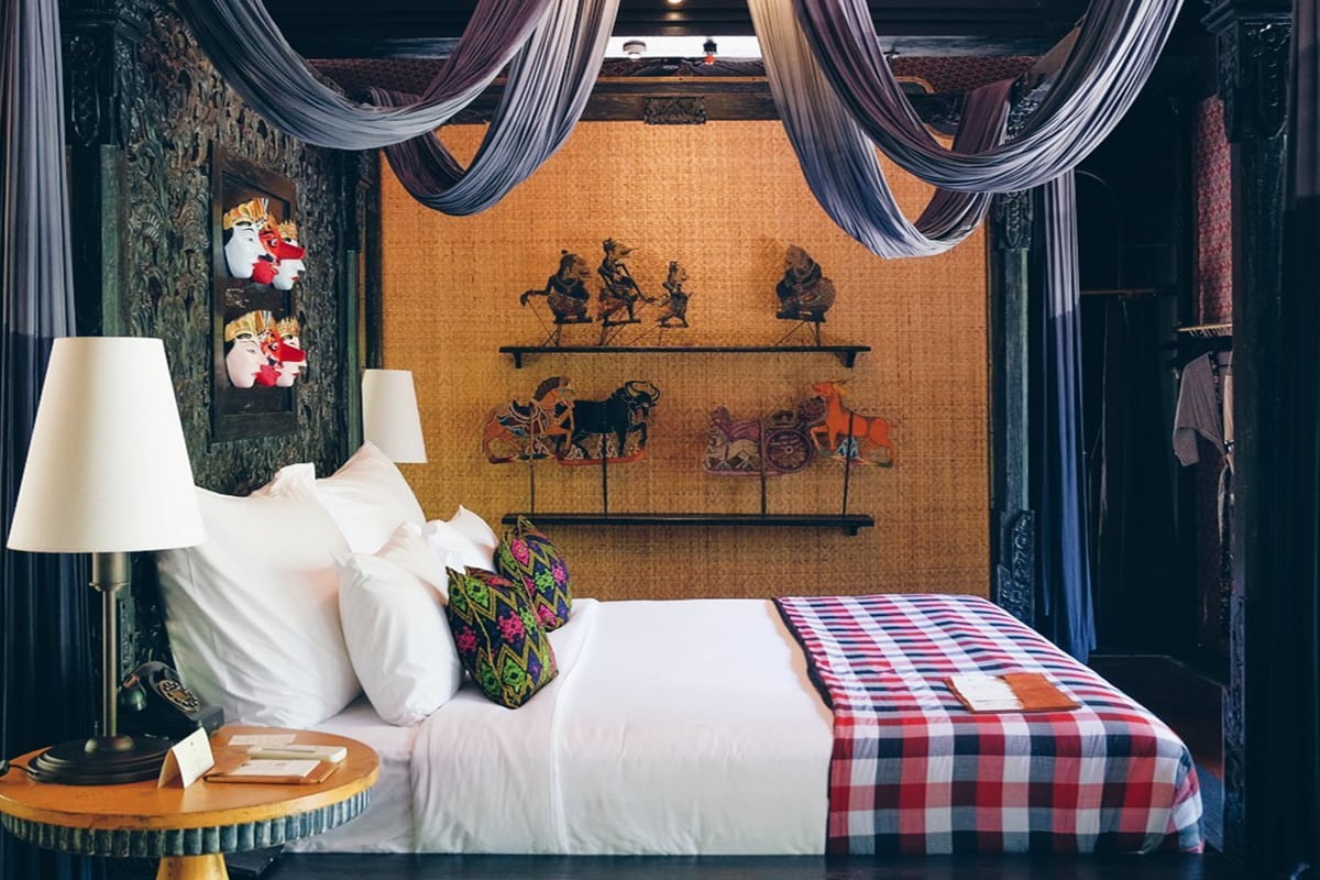 a room inside Capella Ubud, featuring a single big white bed, a plaid blanket, lamp table, and some traditional Indonesian decorations