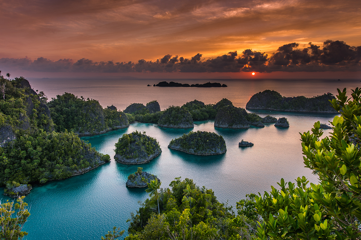 Matahari terbenam di lautan kepulauan Raja Ampat, langit merah jingga, air laut biru gradasi ungu, gugusan pulau bak tempurung kura-kura