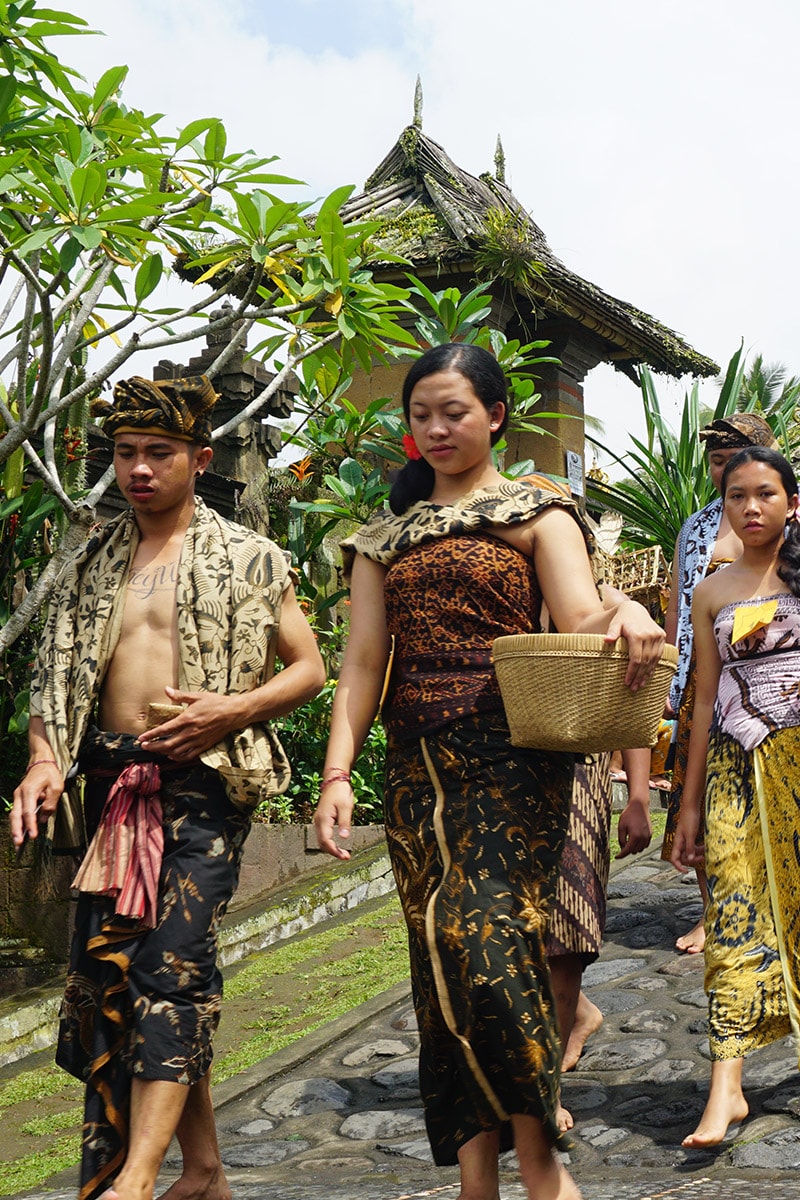 A group of Balinese people in traditional attire walking through a Penglipuran village. A man in the foreground wears a patterned sarong and headpiece, with a woman beside him carrying a woven basket. The architecture features Balinese temple structures with tropical plants surrounding the stone pathway. - Penglipuran Festival 2023